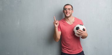 Futbol futbol topu sürpriz bir fikir ya da soru işaret parmağı ile gülen yüz, bir numara tutarak gri grunge duvarın üzerinden beyaz delikanlı