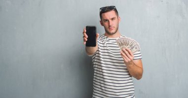 Beyaz delikanlı holding sorunu nedeniyle dolar ve smarphone şüpheci ve sinir, papatyanın üzgün gri grunge duvarın üzerinden. Negatif kişi.