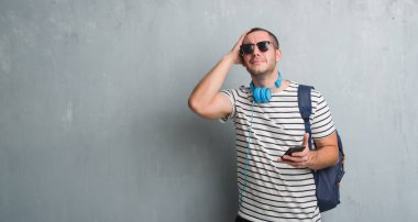 Gri grunge duvar başında, el ile vurguladı smartphone kullanarak kulaklık giyen genç beyaz öğrenci adam şok utanç ve sürpriz suratla, öfkeli ve sinirli. Korku ve hata için üzgün.