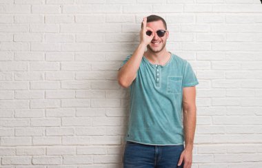 Mutlu yüz el parmaklarının arasından seyir göz üzerinde ok işaretiyle yapıyor gülümseyen güneş gözlüğü takmış beyaz tuğla duvarın üzerinden ayakta beyaz delikanlı