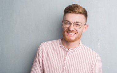 Genç Kızıl saçlı adam ayakta ve diş gösterilen kendine güvenen bir gülümsemeyle gülümseyerek mutlu bir yüz ile gri grunge duvarın üzerinden