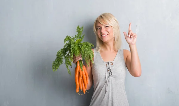 白人成年妇女在灰色的垃圾墙持有新鲜胡萝卜惊讶的想法或问题指着一个快乐的脸 — 图库照片