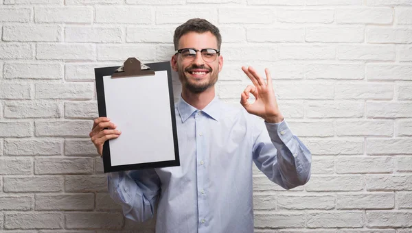優秀なシンボルの指で サインをしているレンガ壁持株クリップボード上の若い大人のビジネス人 — ストック写真