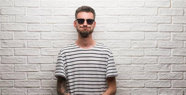 Genç Yetişkin giyen güneş gözlüğü komik surat ile yanakları şişirme beyaz tuğla duvarın üzerinden duran adam. Ağız ile hava, deli ifade şişirilmiş.