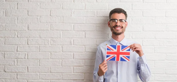 立っていると自信を持って歯を見せて笑顔で笑っている幸せそうな顔でイギリスの旗を保持しているレンガの壁を越えて若い成人男性 — ストック写真