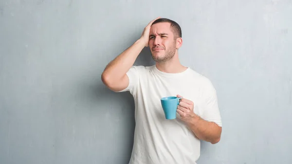 Young caucasian man over grey grunge wall driking a cup of coffee stressed with hand on head, shocked with shame and surprise face, angry and frustrated. Fear and upset for mistake.