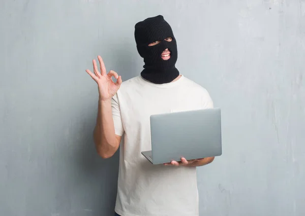 灰色垃圾墙上的年轻白种人黑客做网络攻击使用笔记本电脑做 标志与手指 优秀的符号 — 图库照片