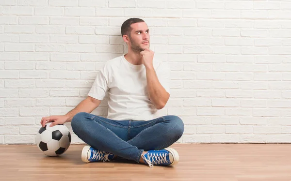 年轻的白种人坐在白色砖墙上拿着足球足球严肃的面孔思考问题 非常困惑的想法 — 图库照片