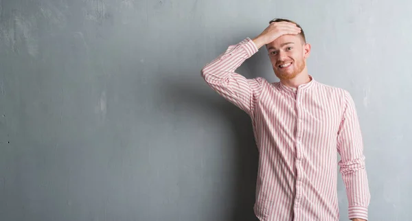 Young redhead man over grey grunge wall stressed with hand on head, shocked with shame and surprise face, angry and frustrated. Fear and upset for mistake.