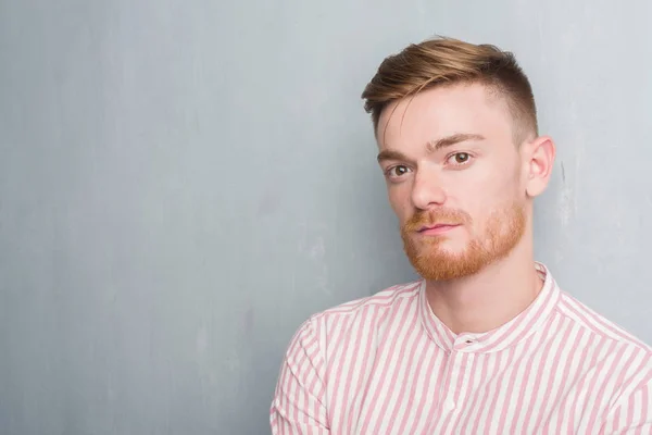 Young redhead man over grey grunge wall with a confident expression on smart face thinking serious