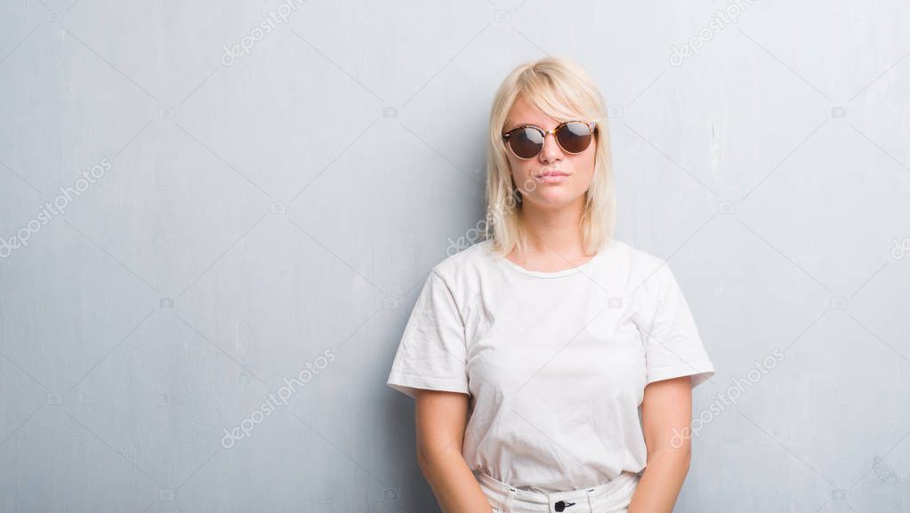 Adult caucasian woman over grunge grey wall wearing sunglasses with serious expression on face. Simple and natural looking at the camera.