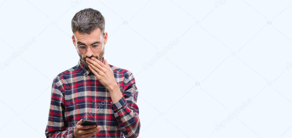 Young hipster adult man holding smartphone cover mouth with hand shocked with shame for mistake, expression of fear, scared in silence, secret concept