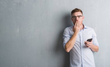 Hata, korku, sessizlik içinde gizli kavramı korkmuş ifade için utanç ile genç Kızıl saçlı adam akıllı telefon kapağı ağız el ile kullanarak ileti gönderme gri grunge duvarın üzerinden şok