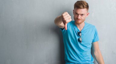 Genç Kızıl saçlı adam mutsuz ve kızgın ret gösterilen ve başparmak aşağı hareketi ile negatif seyir gündelik kıyafeti giymiş gri grunge duvarın üzerinden. Kötü ifade.