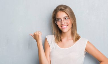 İşaret ve başparmak kadar mutlu yüz gülümseyen tarafı ile gösteren gözlük takan grunge gri duvar üzerinde duran güzel genç kadın