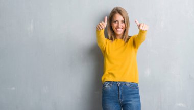 Grunge gri duvar onaylama üzerinde duran güzel genç kadın el ile pozitif jest yapıyor gülümseyen ve başarı için mutlu yaşasın. Kamera, kazanan jest arıyorsunuz.