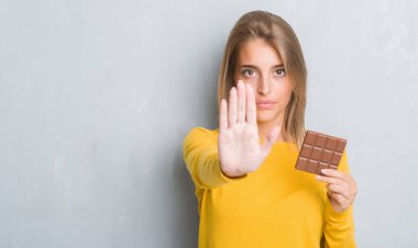 Grunge gri duvar yeme üzerinde güzel genç kadın çikolata bar açık eliyle dur işareti ile ciddi ve kendine güvenen ifade, savunma hareketi yapıyor