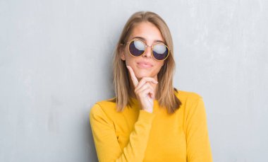 Retro güneş gözlüğü ciddi bir yüz düşünme hakkında soru, çok karışık fikir giyen grunge gri duvar üzerinde duran güzel genç kadın