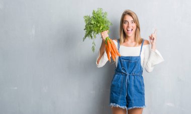 Grunge gri güzel genç kadın taze havuç sürpriz bir fikir ile tutarak duvar veya gülen yüz, bir numara ile işaret parmağı soru