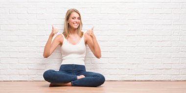 El ile pozitif jest yapıyor başarı işareti yere evde oturmak güzel ve genç birbayan başparmak gülümseyen ve mutlu. Neşeli bir ifadeyle, kazanan jest kameraya bakıyor.