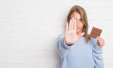 Güzel genç kadın çikolata ile yemek beyaz tuğla duvarın üzerinden açık el dur işareti ile ciddi ve kendine güvenen ifade, savunma hareketi yapıyor