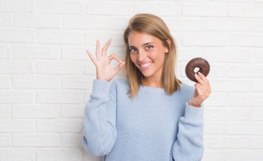 Güzel genç kadın parmakları, mükemmel sembolü olan ok işareti yapıyor çikolatalı çörek yemek beyaz tuğla duvarın üzerinden