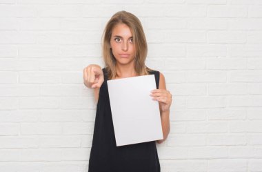 Güzel genç kadın kamera ve size parmak ile işaret boş kağıt sayfası holding beyaz tuğla duvarın üzerinden, el işareti, pozitif ve kendinden emin bir jest ön