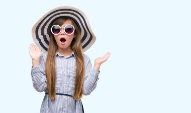 Şapka ve güneş gözlüğü çok mutlu ve heyecanlı, giyen genç sarışın bebek kazanan ifade büyük gülümseme ile çığlık ve ellerini kaldırdı zaferini kutluyor