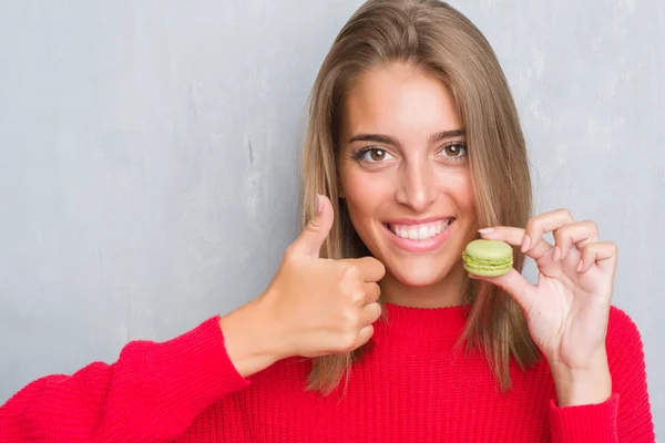 美丽的少妇在垃圾灰色墙壁吃绿色 Macaron 愉快以大微笑做 拇指用手指 优秀标志 — 图库照片