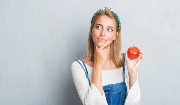 美丽的少妇在垃圾灰色墙壁吃新鲜的蕃茄严肃的面孔思考问题 非常迷惑想法 — 图库照片
