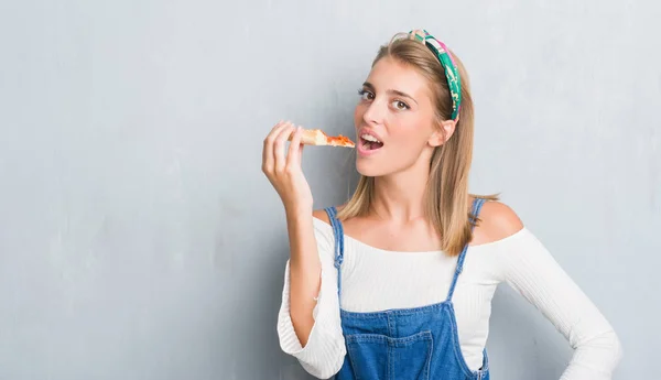 Beautiful young woman over grunge grey wall eating pepperoni pizza slice with a confident expression on smart face thinking serious