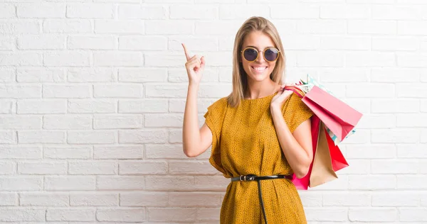 美丽的年轻妇女在白色砖墙举行购物袋在销售惊奇以想法或问题指向手指以愉快的面孔 — 图库照片
