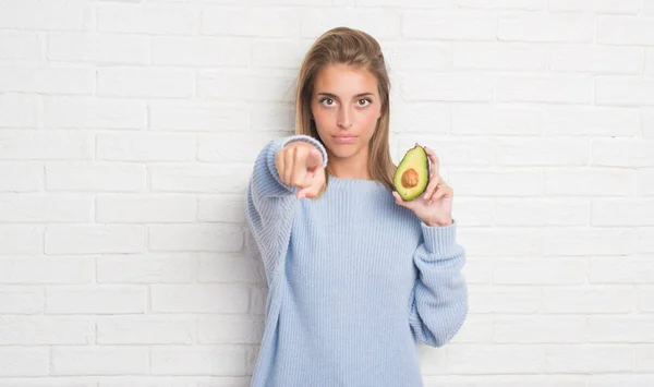 新鮮なアボカドを指す指でカメラと食べる白いレンガの壁に美しい若い女性 手に署名 正面からポジティブで自信のジェスチャー — ストック写真