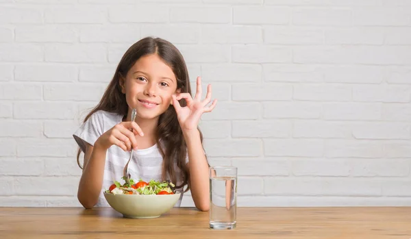 優秀なシンボルの指で サインをしている健康的なサラダを食べるテーブルの上に座って若いヒスパニックの子供 — ストック写真