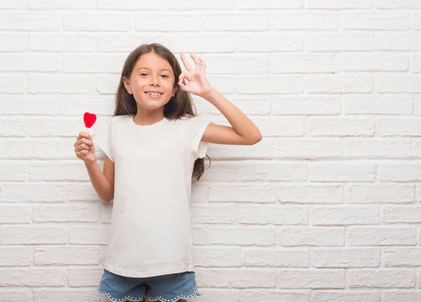 優秀なシンボルの指で サインをしている赤いハート棒付きキャンディーを食べる白いレンガ壁にヒスパニック系の子供 — ストック写真