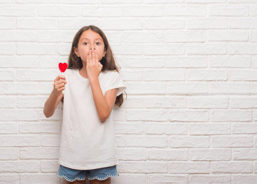 Young hispanic kid over white brick wall eating red heart lollipop candy cover mouth with hand shocked with shame for mistake, expression of fear, scared in silence, secret concept