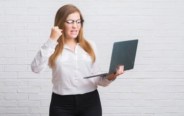 年轻的商业妇女在白色砖墙拿着电脑膝上型的愤怒和沮丧的呼喊 疯狂和咆哮与举手 愤怒的概念 — 图库照片