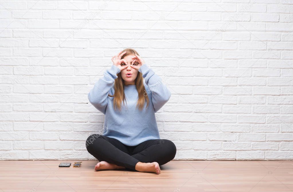 Young adult woman sitting on the floor in autumn over white brick wall doing ok gesture like binoculars sticking tongue out, eyes looking through fingers. Crazy expression.