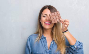 Gri grunge duvar yeme üzerinde Genç Yetişkin kadın ayakta ve diş gösterilen kendine güvenen bir gülümsemeyle gülümseyerek mutlu bir yüz ile çikolata bar