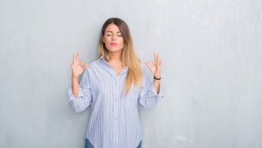 Genç Yetişkin kadın moda iş kıyafeti giymiş gri grunge duvarın üzerinden rahatla ve gülümseyen gözlerle kapalı parmak jestle meditasyon yapıyor. Yoga kavramı.