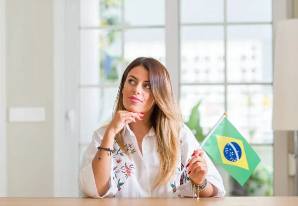 Young woman at home holding flag of Brazil serious face thinking about question, very confused idea