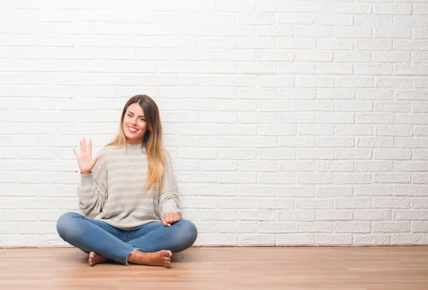 年轻的成年女子坐在地板上的白色砖墙在家里显示和指向五的手指 而微笑着自信和快乐 — 图库照片