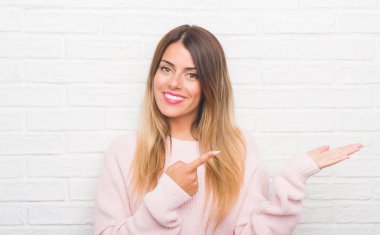 Genç Yetişkin kadın yüzünden beyaz tuğla duvar adlı aşınma kış kıyafeti eve hayran ve el ile sunulması ve parmak ile işaret ederken kameraya gülümseyen.