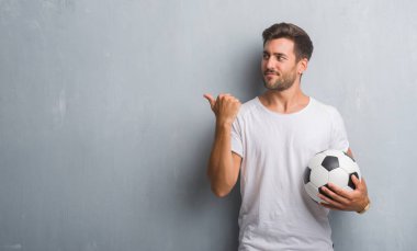 İşaret ve başparmak kadar mutlu yüz gülümseyen tarafı ile gösterilen futbol futbol topu tutan gri grunge duvarın üzerinden yakışıklı genç