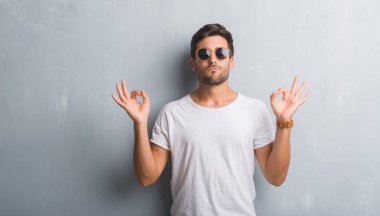 Yakışıklı genç güneş gözlüğü takıyor gri grunge duvarın üzerinden rahatla ve gülümseyen gözlerle kapalı parmak jestle meditasyon yapıyor. Yoga kavramı.