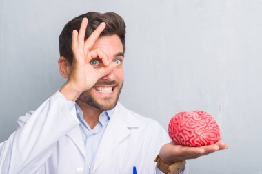 Yakışıklı genç doktor psikolog adam bir beyin ile ok işareti ile el parmaklarının arasından seyir göze yapıyor gülümseyen mutlu yüz tutan gri grunge duvarın üzerinden