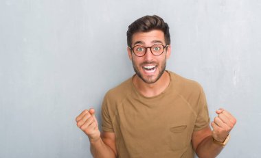 Yakışıklı delikanlı kutluyor gözlük takan gri grunge duvarın üzerinden şaşırttı ve başarı için kaldırdı kollarını açarak hayret ve gözleri açık. Kazanan kavramı.
