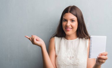 Grunge genç esmer kadın defter çok mutlu el ve parmak ile tarafına işaret gösterilen duvar gri