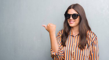 Genç esmer kadın işaret ve başparmak kadar mutlu yüz gülümseyen tarafı ile gösterilen büyük güneş gözlüğü takıyor grunge gri duvarın üzerinden