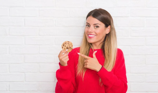Young adult woman over white brick wall eating chocolate chip cooky very happy pointing with hand and finger